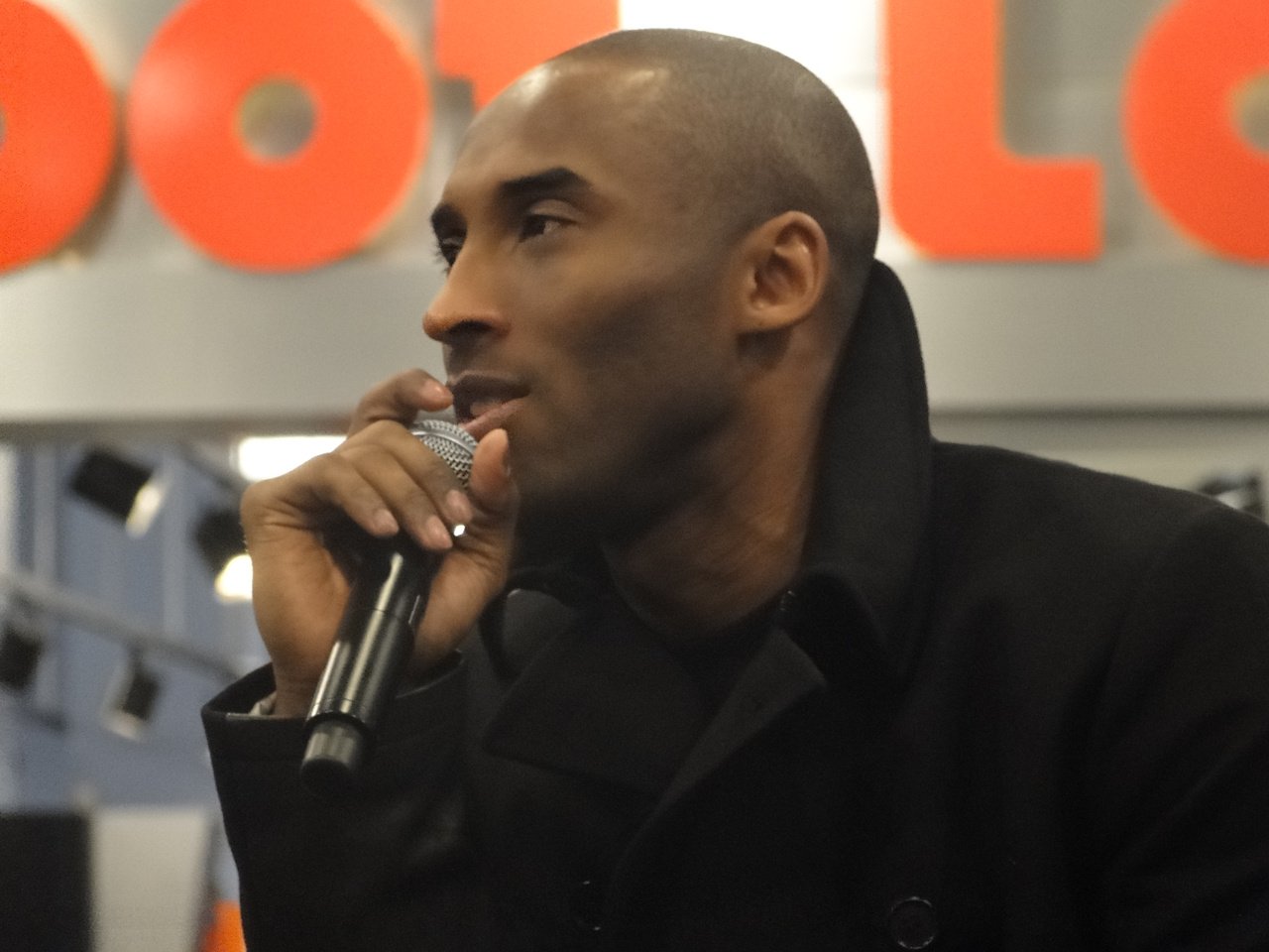 Kobe Bryant holds a microphone and looks to his right, wearing a black jacket.